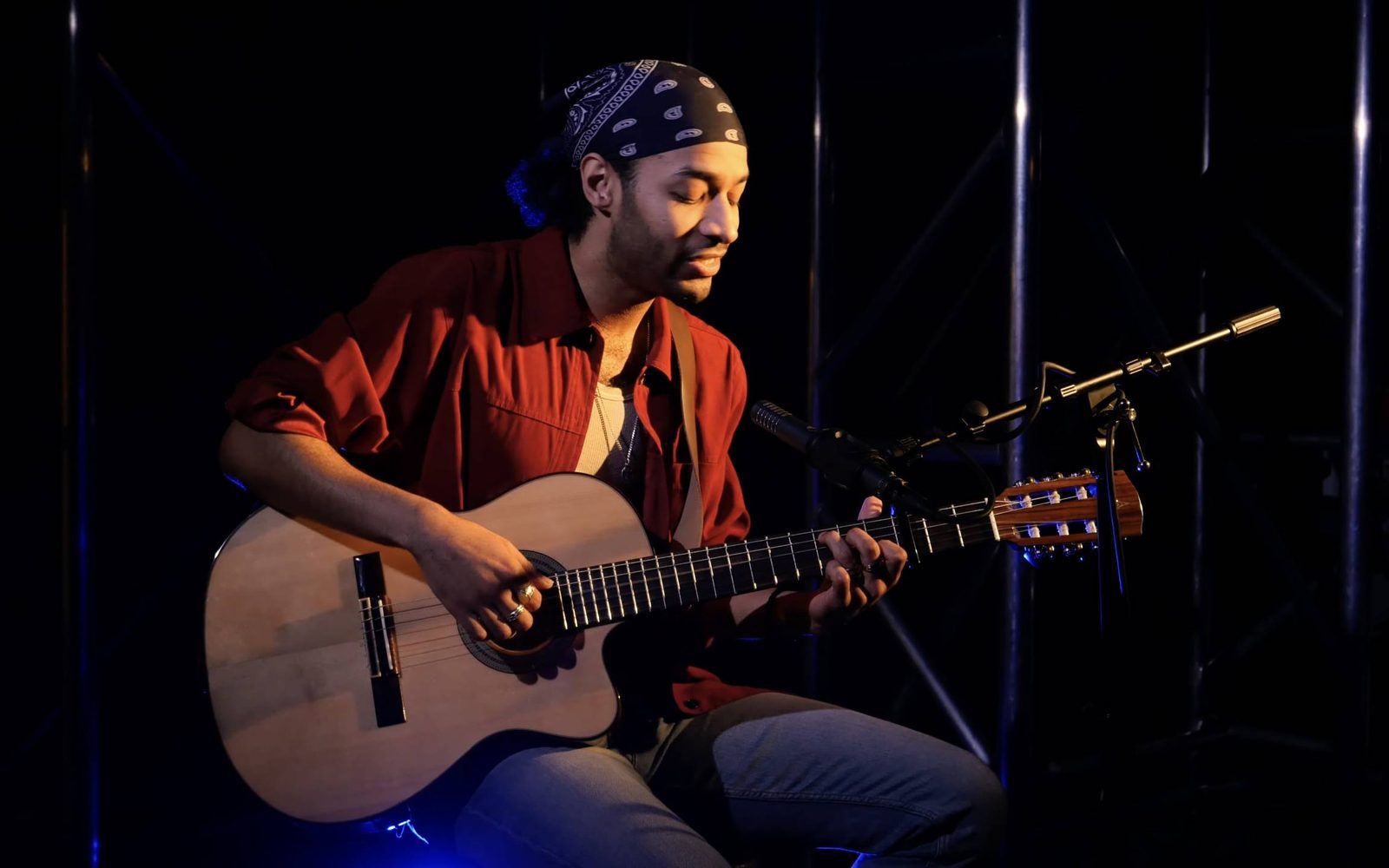Jordan Dobson in a black bandana and red button-down sits in a darkened room playing guitar.